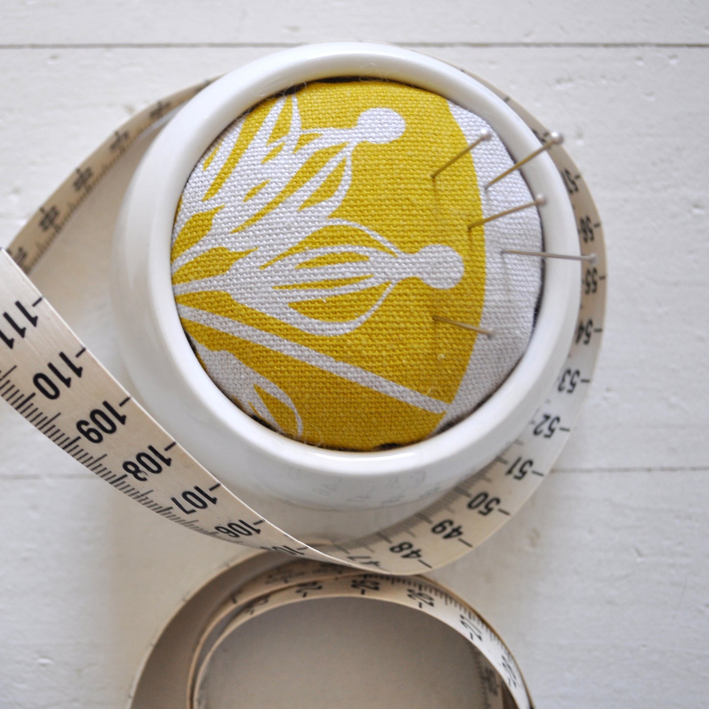 Pinch River ceramic pin cushion featuring Firewheel print in Buddha Gold (colourway 1_v2). Close-up top view. Product image is styled with a measuring tape and pins.