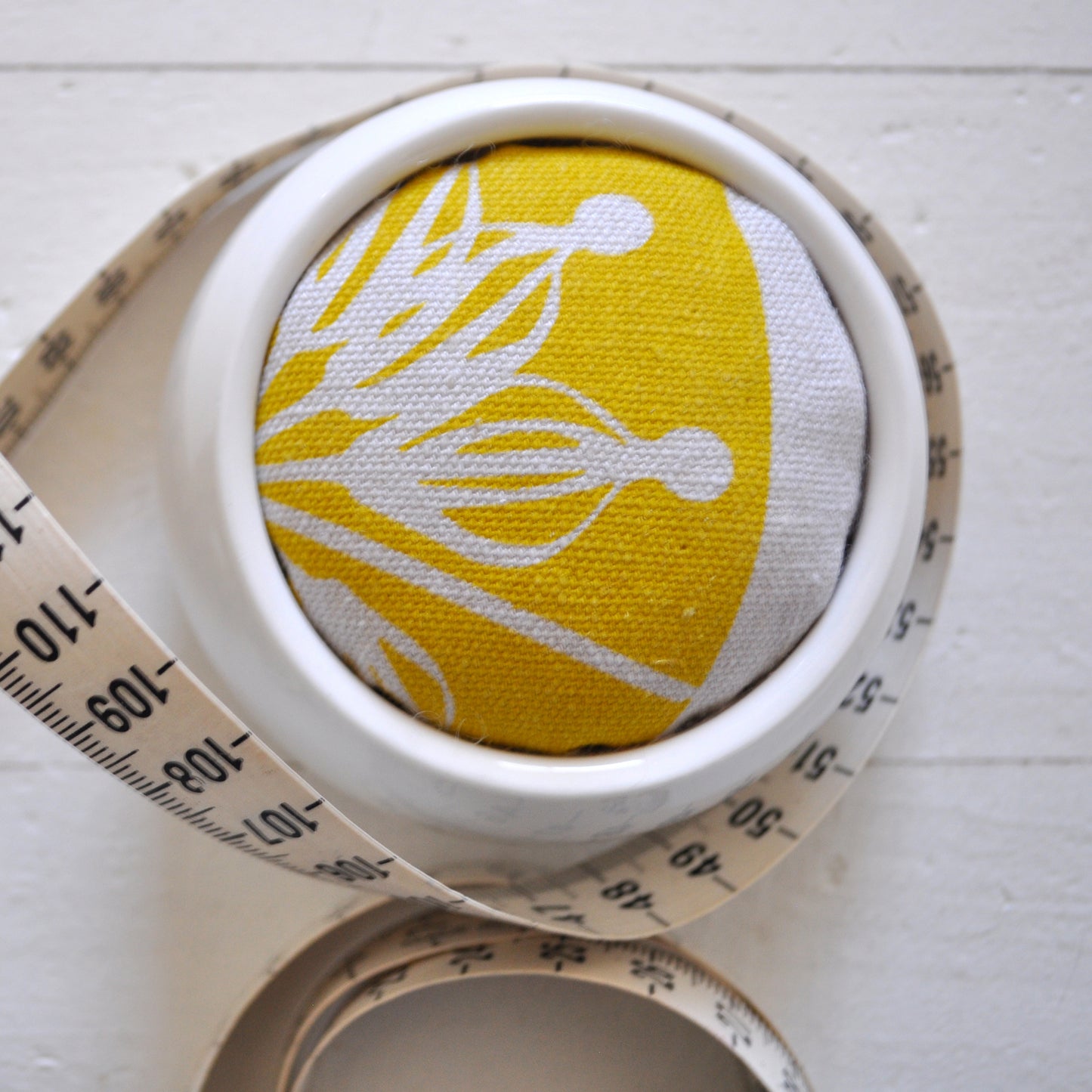 Pinch River ceramic pin cushion featuring Firewheel print in Buddha Gold (colourway 1_v2). Close-up top view. Product image is styled with a measuring tape.