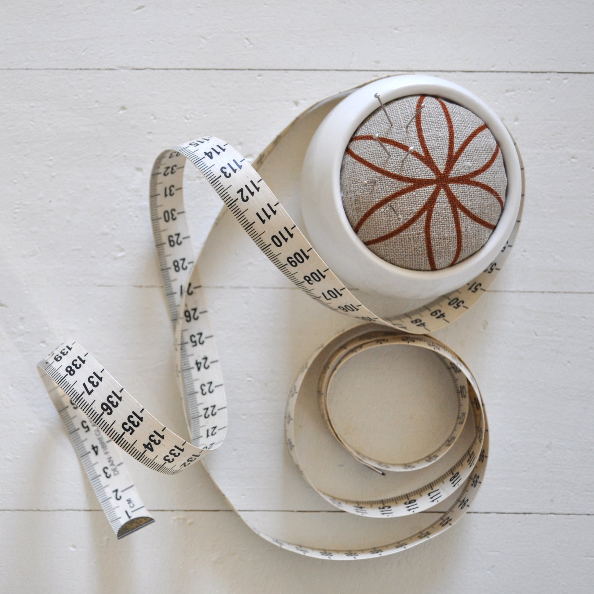Pinch River ceramic pin cushion featuring Flower of Life print in Burnt Orange (colourway 2). Top view. Product image is styled with a measuring tape and pins.