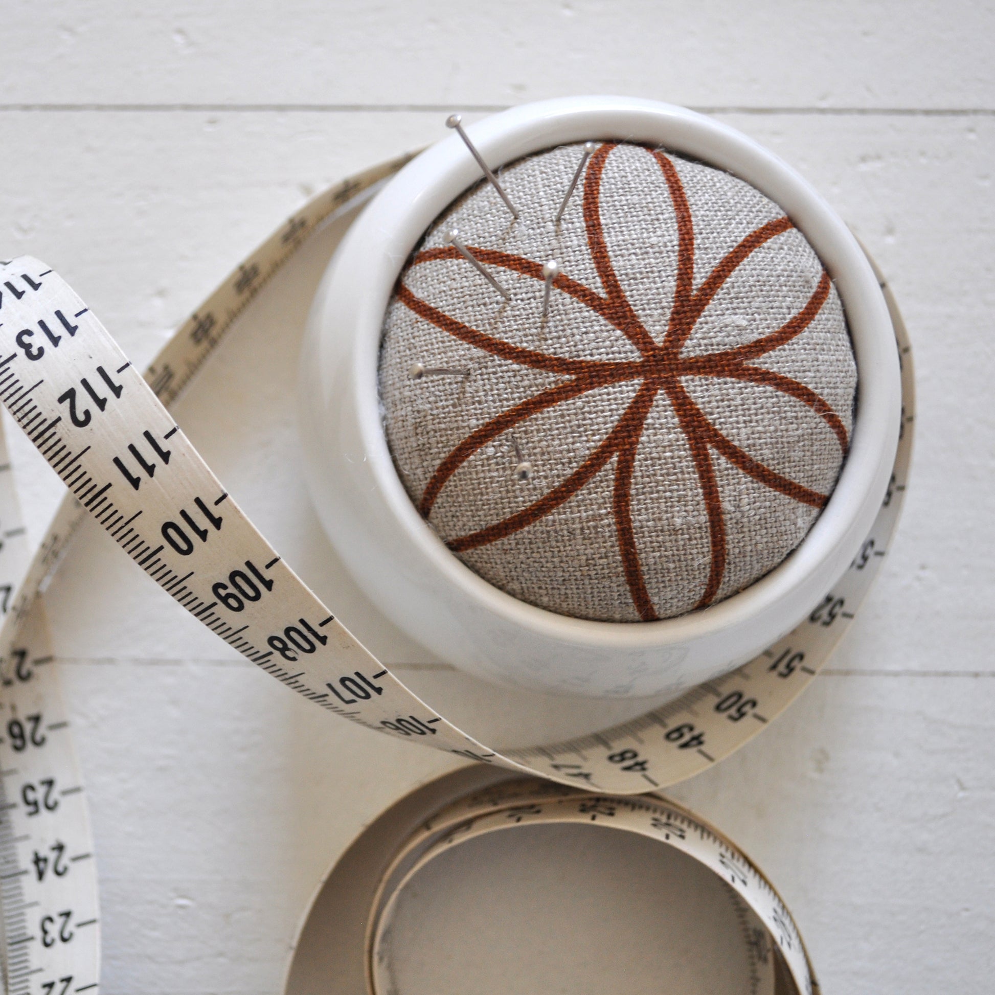 Pinch River ceramic pin cushion featuring Flower of Life in Burnt Orange (colourway 2). Close-up top view. Product image is styled with a measuring tape and pins.