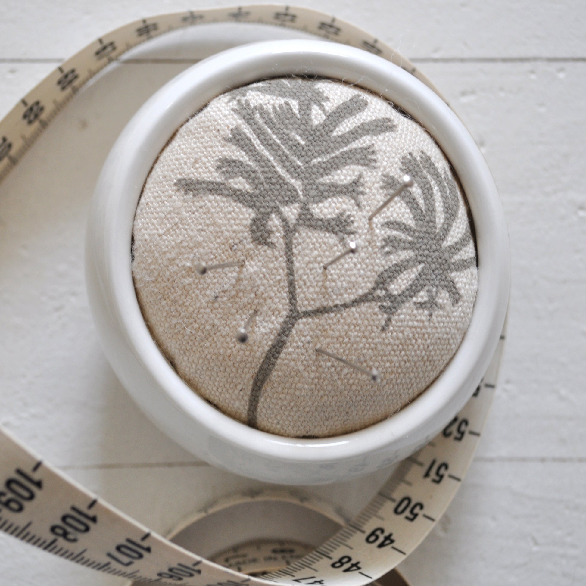 Pinch River ceramic pin cushion featuring Kangaroo Paw print in Aged Wood (colourway 2_v1). Close-up top view. Product image is styled with a measuring tape and pins.