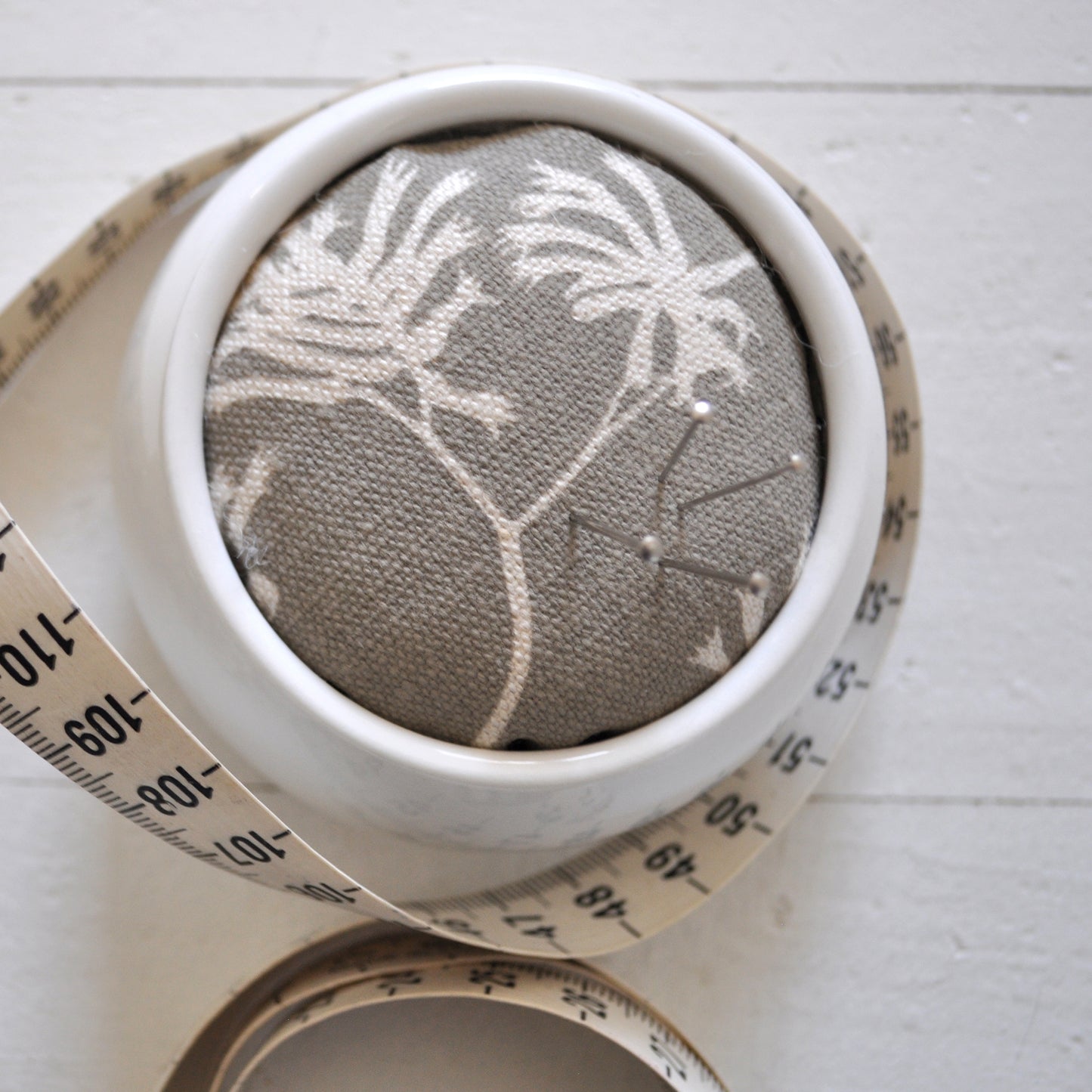 Pinch River ceramic pin cushion featuring Kangaroo Paw print in Aged Wood (colourway 2_v2). Close-up top view. Product image is styled with a measuring tape and pins.