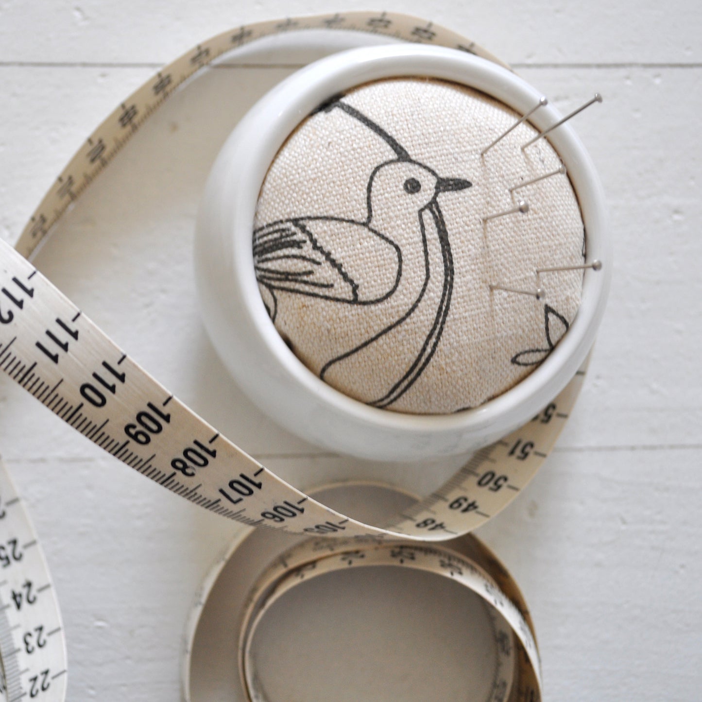 Pinch River ceramic pin cushion featuring Lyrebird print in Grey Bark (colourway 1). Close-up top view. Product image is styled with a measuring tape and pins.