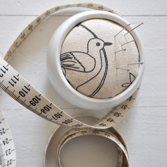 Pinch River ceramic pin cushion featuring Lyrebird print in Grey Bark (colourway 1). Close-up top view. Product image is styled with a measuring tape and pins.