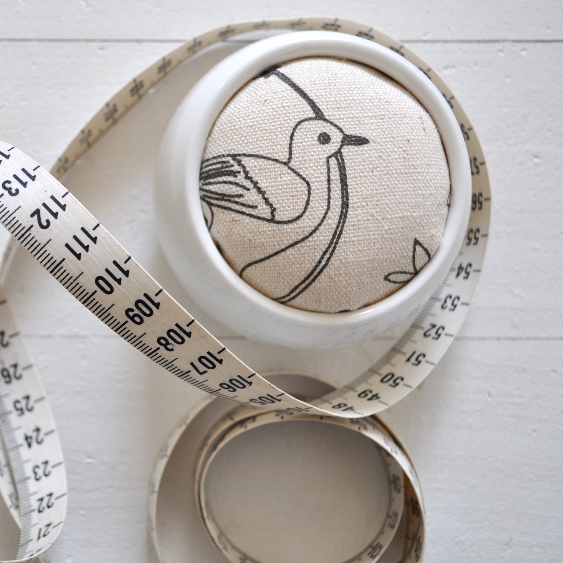 Pinch River ceramic pin cushion featuring Lyrebird print in Grey Bark (colourway 1). Close-up top view. Product image is styled with a measuring tape.
