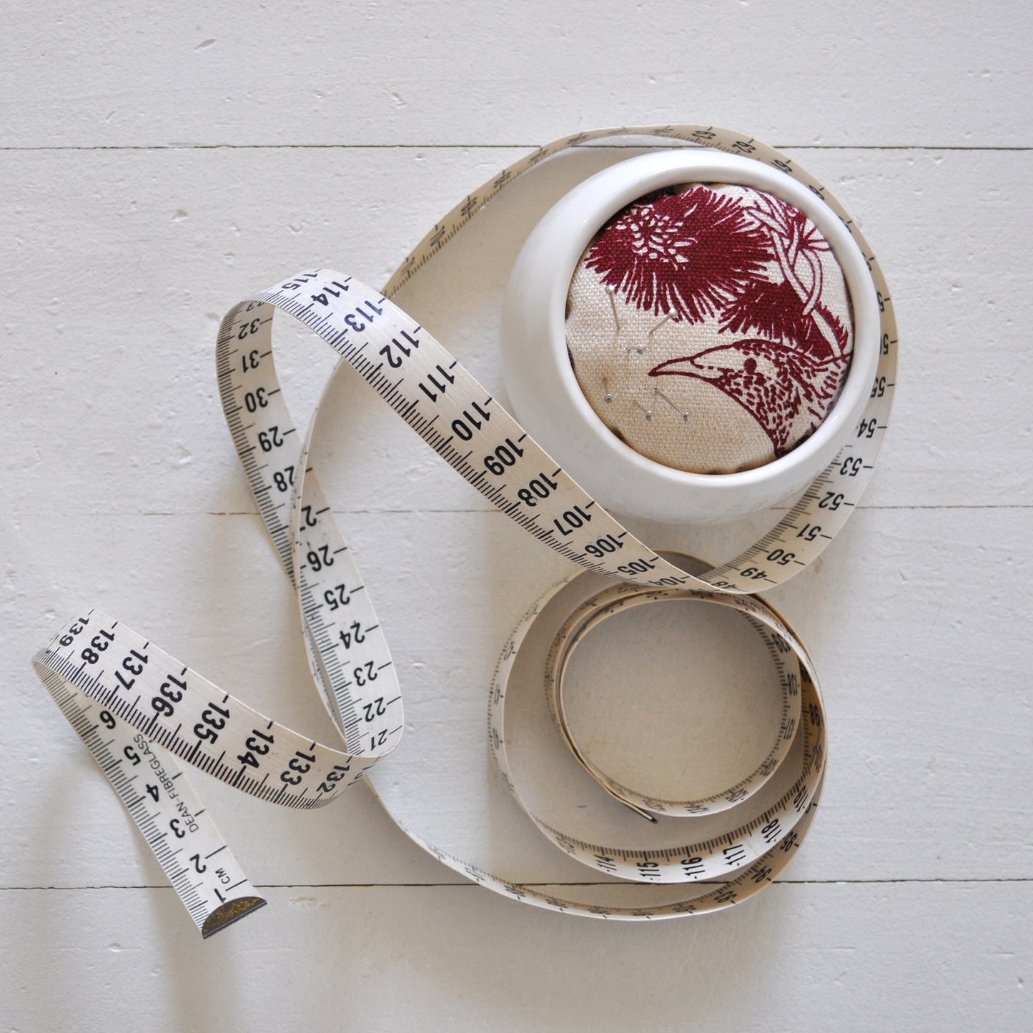Pinch River ceramic pin cushion featuring Wattle Bird print in Wine Berry (colourway 1). Top view. Product image is styled with a measuring tape and pins.