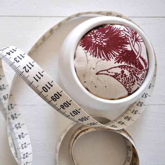 Pinch River ceramic pin cushion featuring Wattle Bird print in Wine Berry (colourway 1). Close-up top view. Product image is styled with a measuring tape and pins.