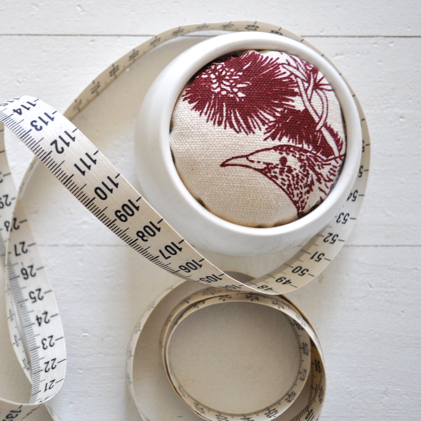 Pinch River ceramic pin cushion featuring Wattle Bird print in Wine Berry (colourway 1). Close-up top view. Product image is styled with a measuring tape.