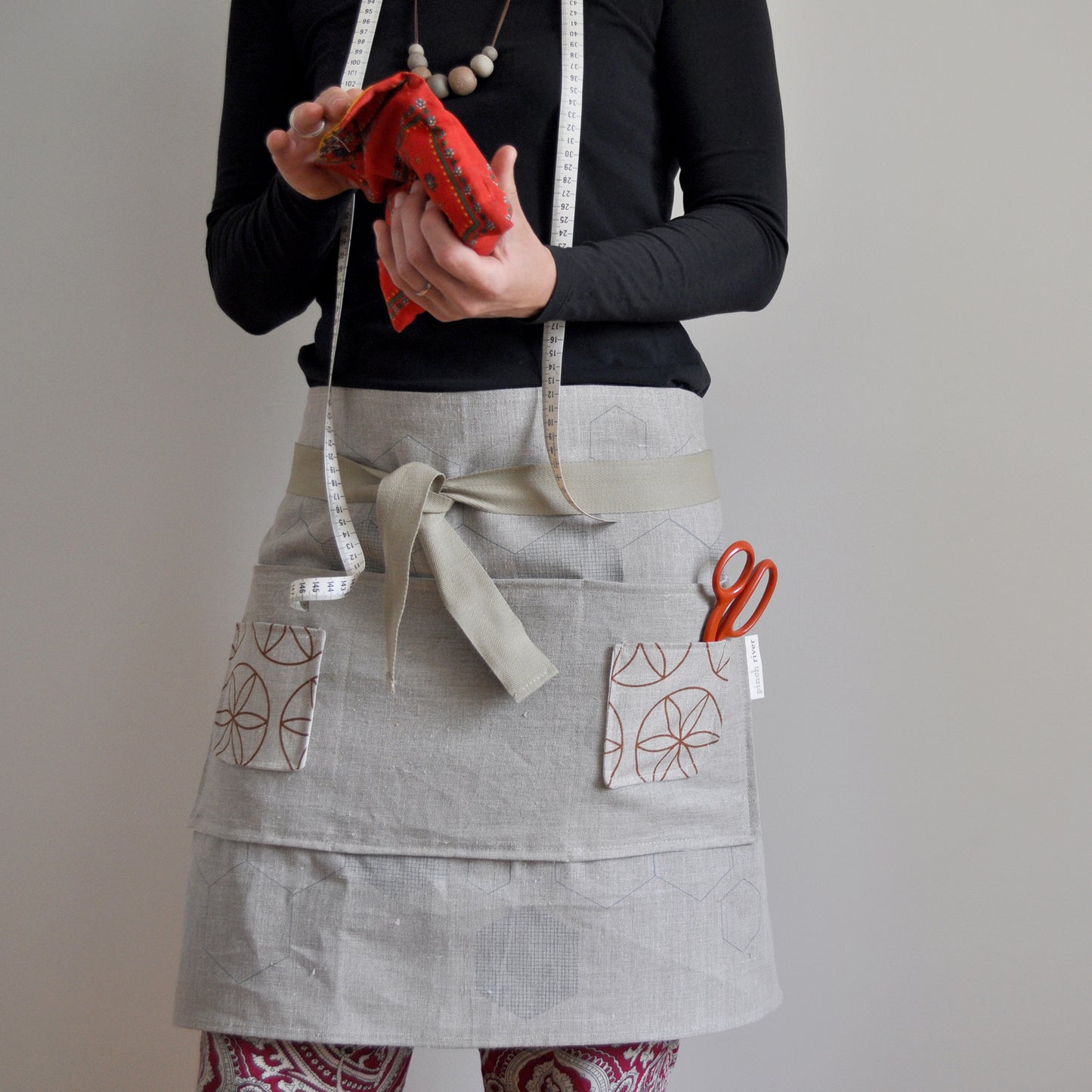 Model wearing Pinch River waist apron / half apron #2, with scissors in pocket, holding a sewing pouch with a measuring tape around her neck, against a plain background. Front view.