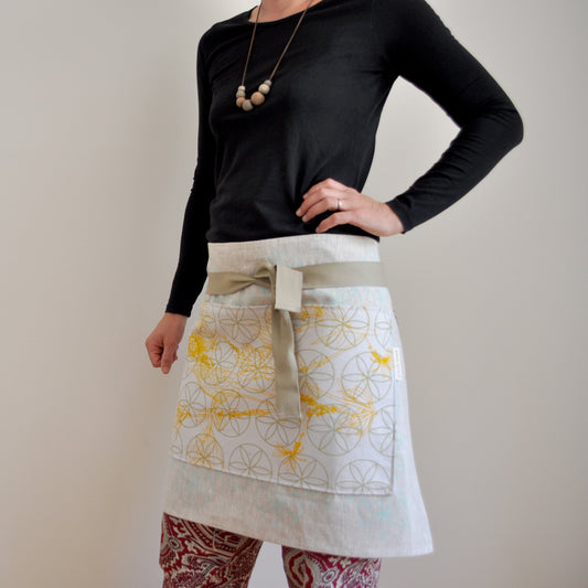 Model wearing Pinch River waist apron / half apron No. 3, holding a potted plant and gardening tools against a plain background. Side View.