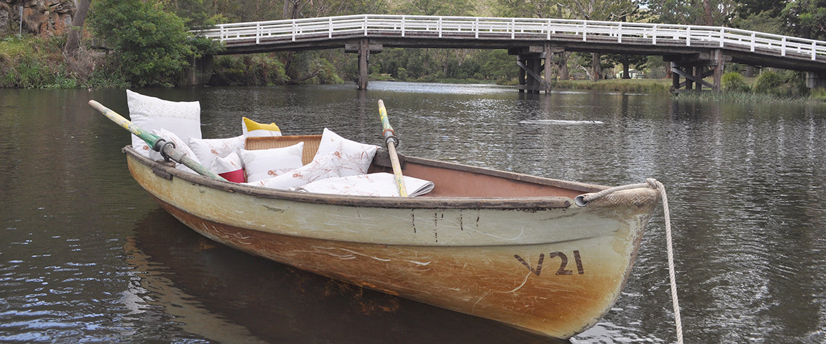 Rowboat on a river with a bridge in the background. Pinch River cushions and quilted throw in the row boat.