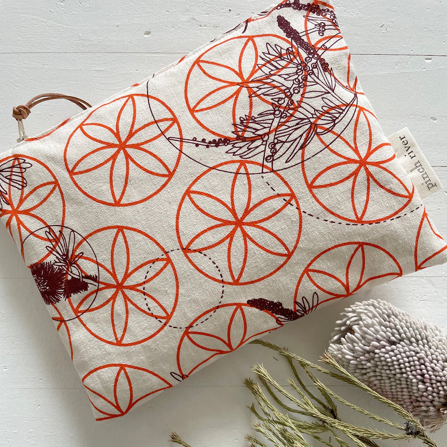 Pinch River zipper pouch featuring Wattle Bird in the Bottle Brush print in Wine Berry (colourway 1) overprinted with Flower of Life print in Fiery Orange (colourway 3). Top view. Product image is styled with Australian native flora.