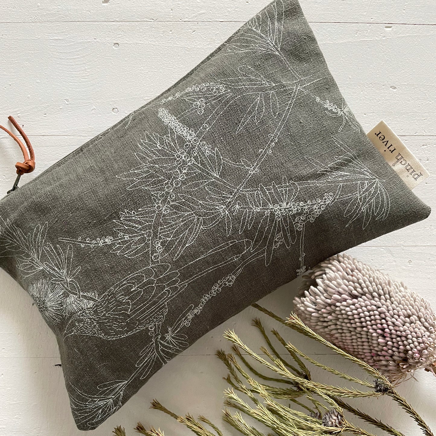 Pinch River zipper pouch featuring Wattle Bird print in Moss Green (colourway 3). Top view. Product image is styled with Australian native flora.